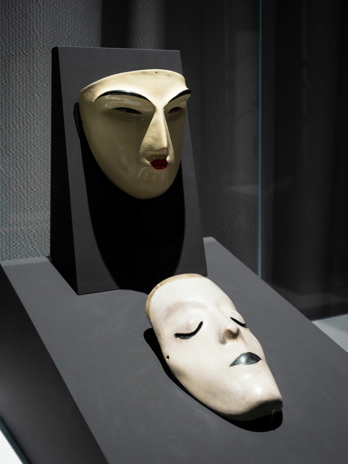 Two white masks with strongly drawn contours around the eyes and mouth in a display case - one hanging, the other lying down.