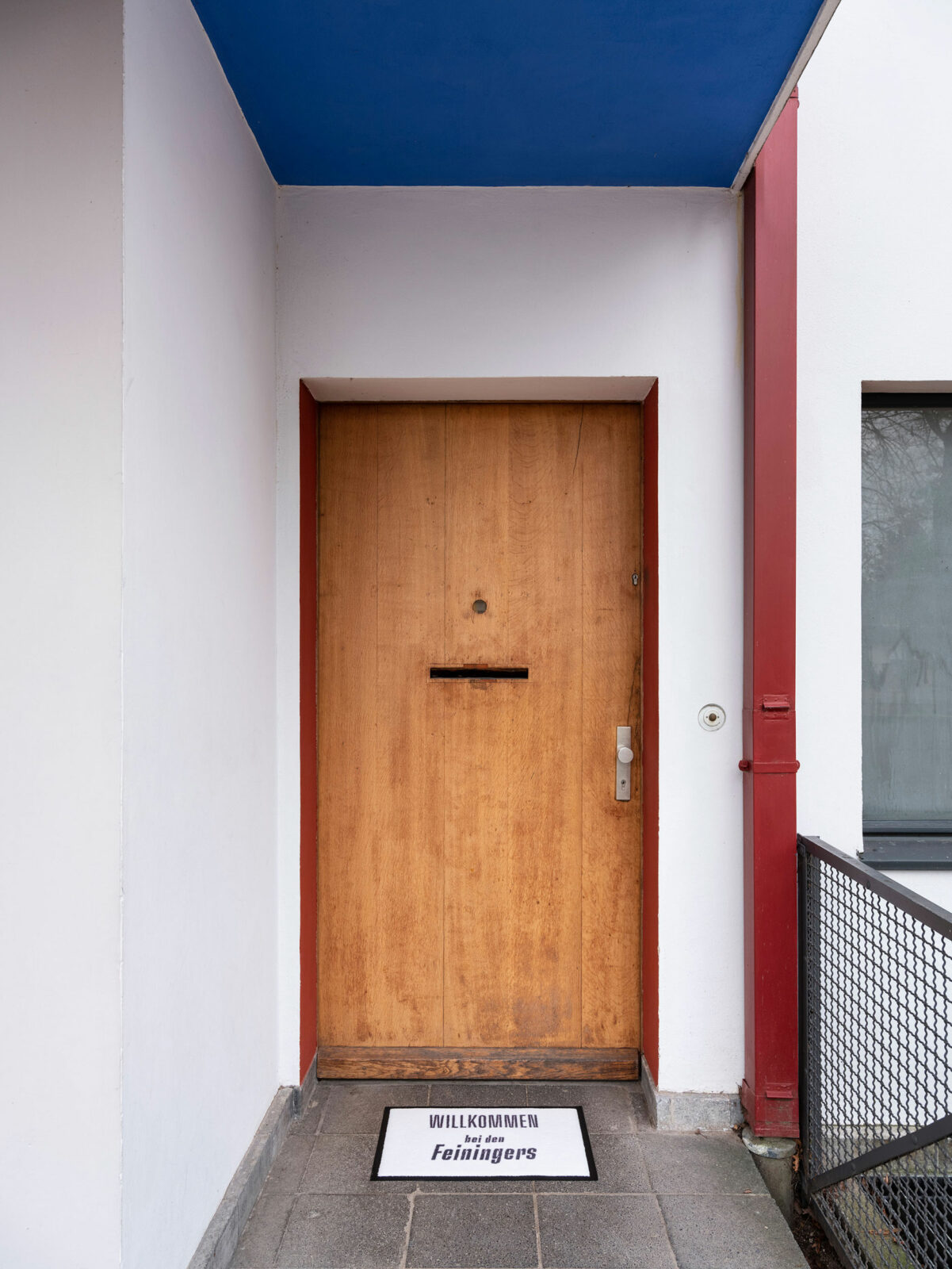 Wooden entrance door with red frame. The ceiling of the cantilevered upper floor is painted blue. In front of the door is a doormat with the inscription 