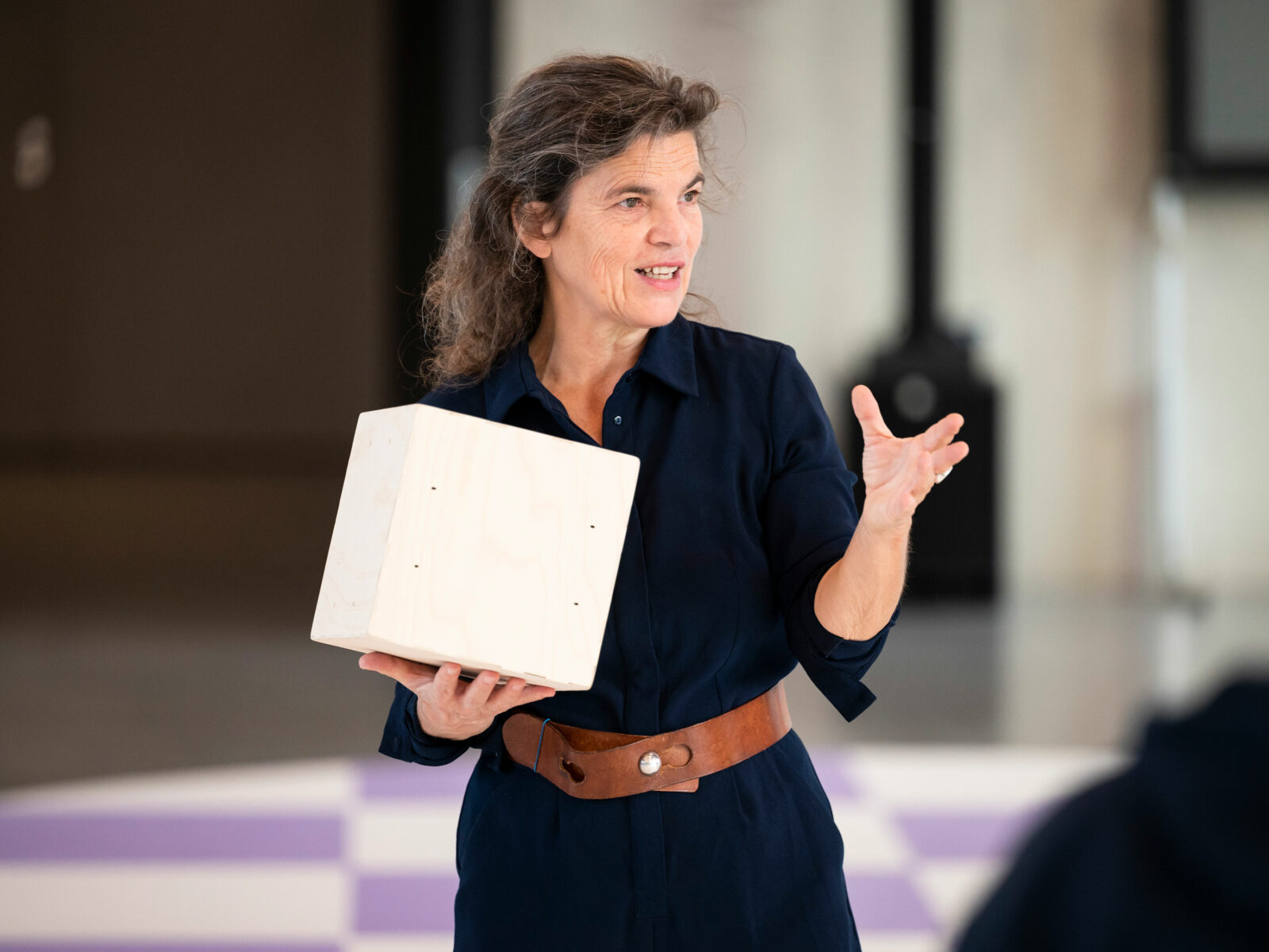 A woman holds a large cube in one hand and gestures with the other.