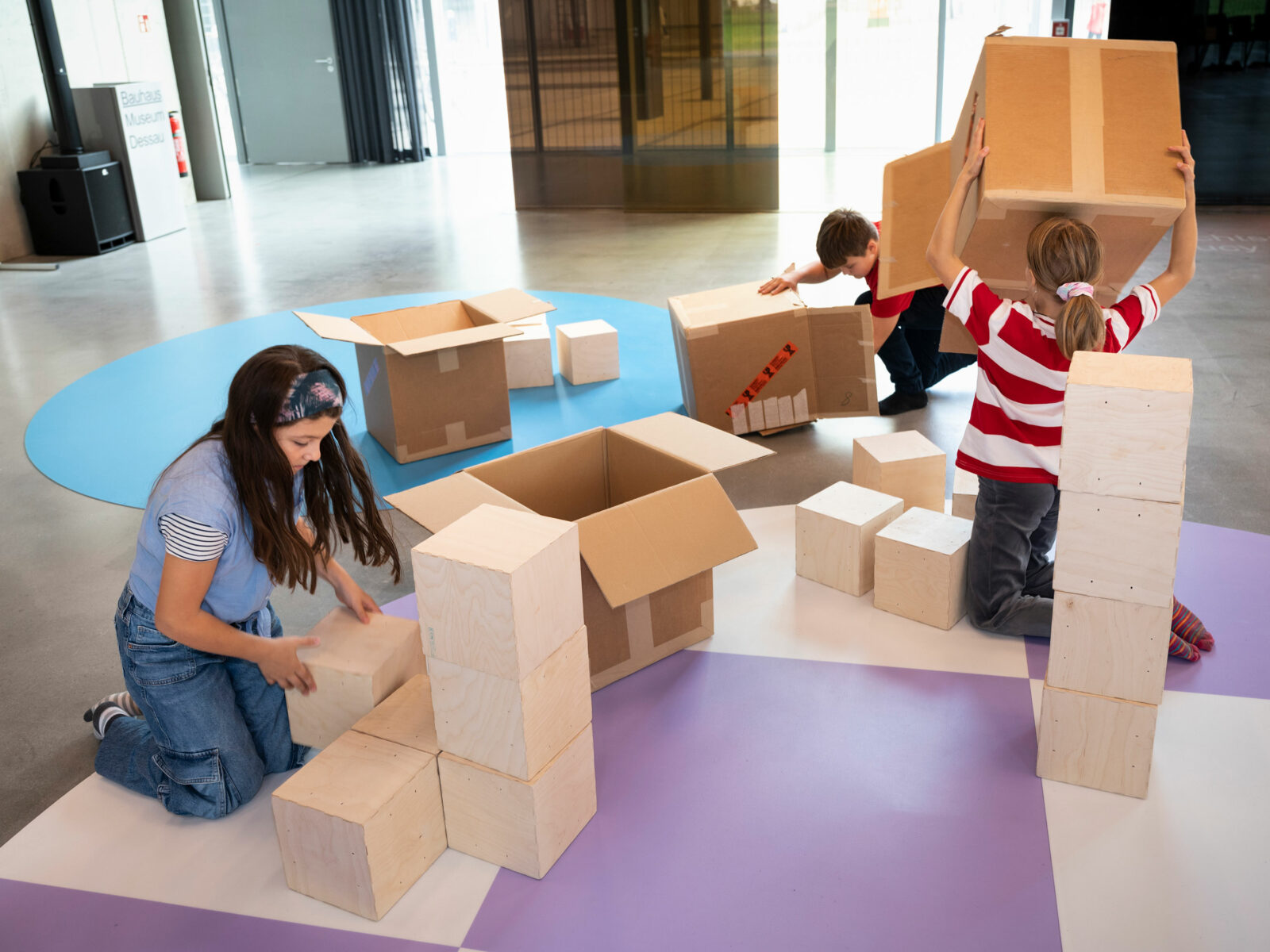 Three children stack boxes and cubes on top of each other. They kneel on the ground.