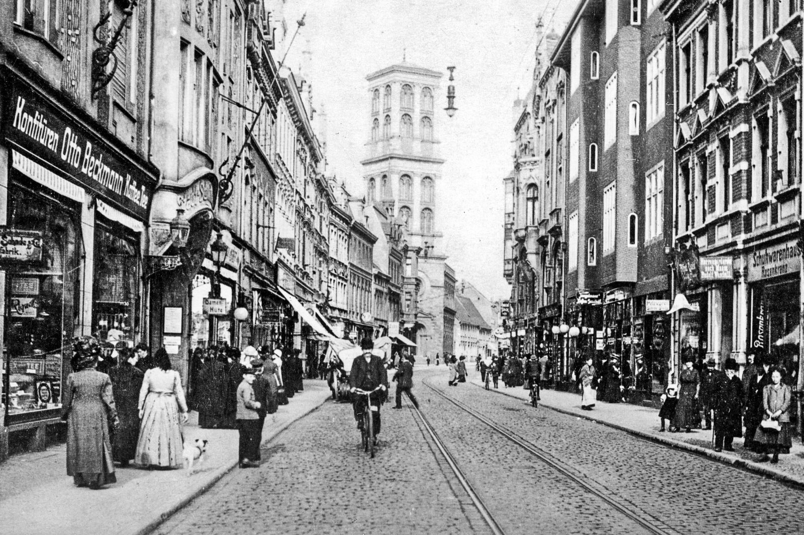 View of a street in the historic residential town of Dessau. On the pavement, women in long dresses walk past stately shops. A cyclist approaches on the street.