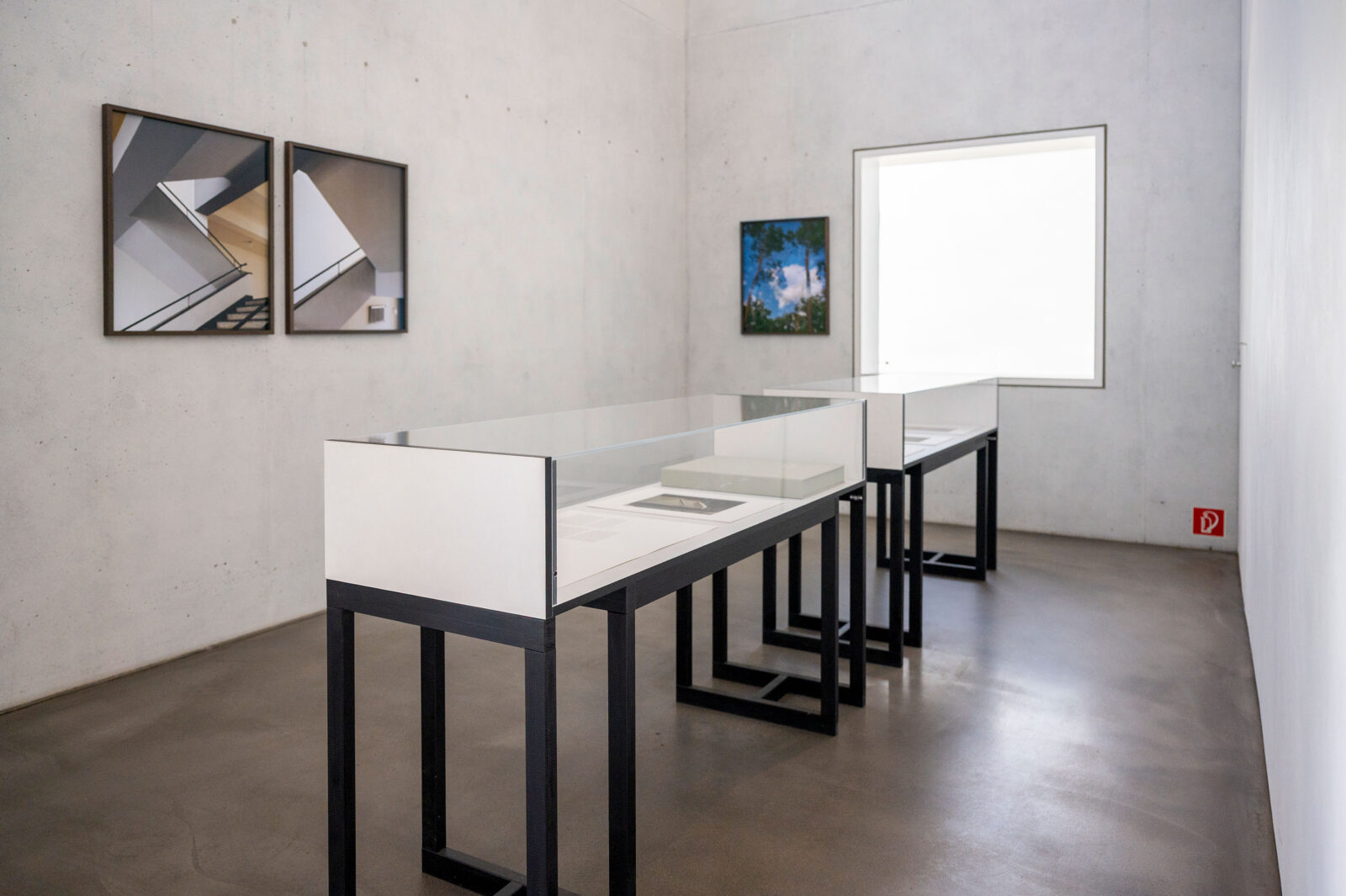Ausstellungsraum mit zwei Vitrinen und 3 Fotografien von Margret Hoppe, die an der Wand hängen. Durch das milchige Fenster des Haus Gropius dringt weiches Licht in den Raum.