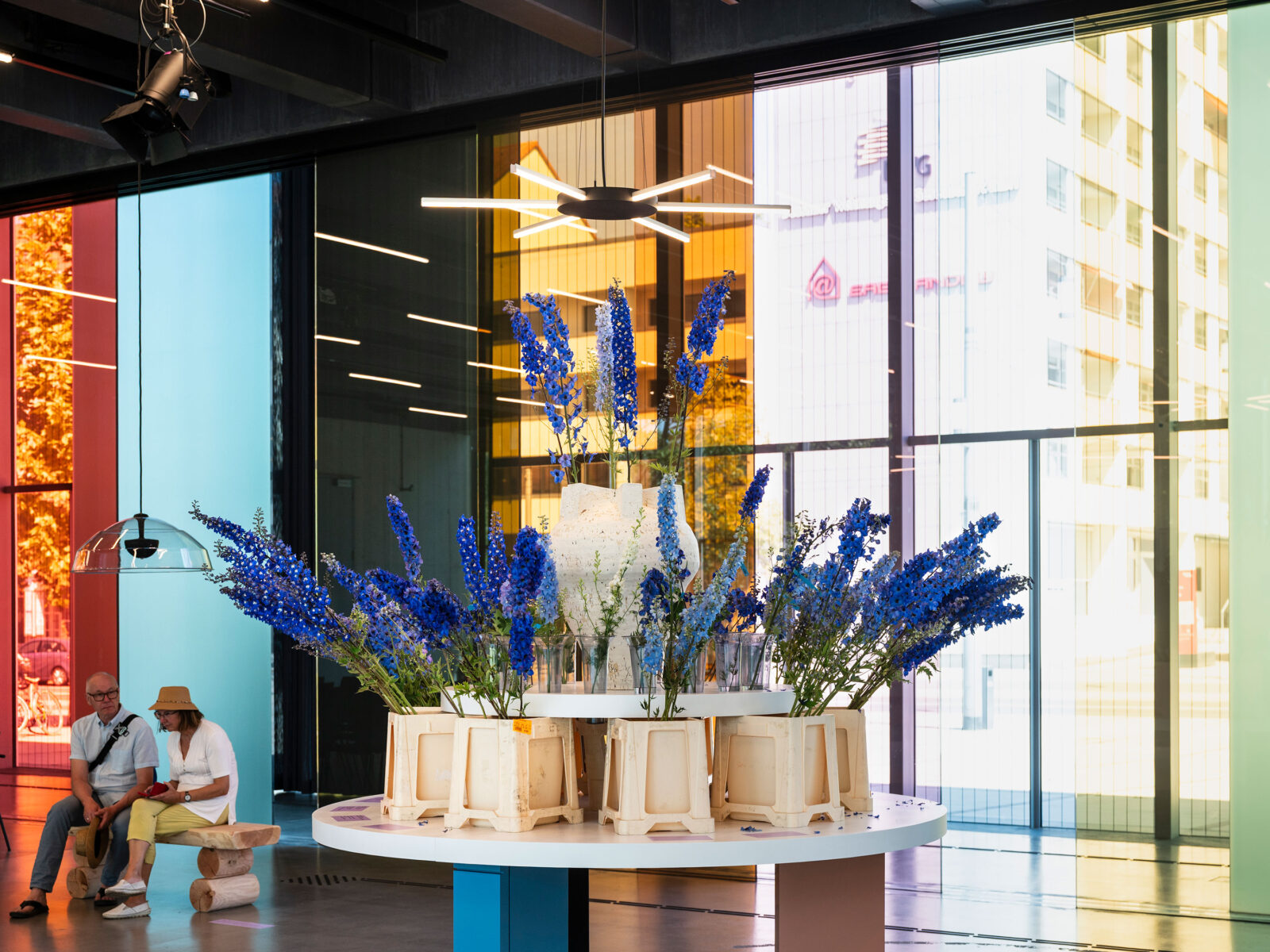 Im Vordergrund ein runder Tisch mit vielen Ritterspornsträußen. In der Mitte eine große Vase mit einzelnen Blütenständen. Im Hintergrund sieht mean Besucher auf einer Bank, die der Sounddusche lauschen, die über dieser hängt.